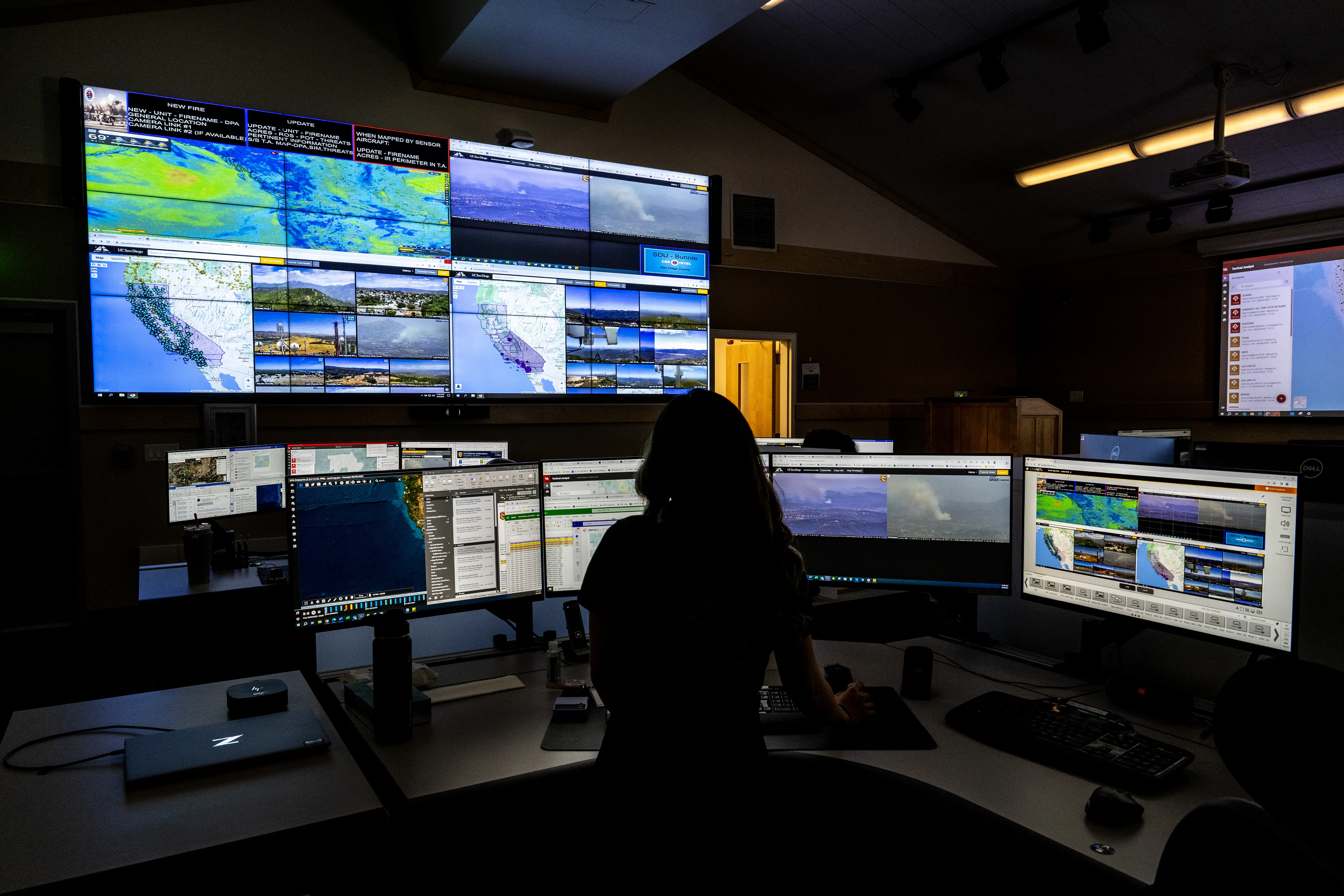 Dark command center with a wall of large, tiled computer screens showing maps and weather radar. A person sits at a desk with multiple monitors in front of them.