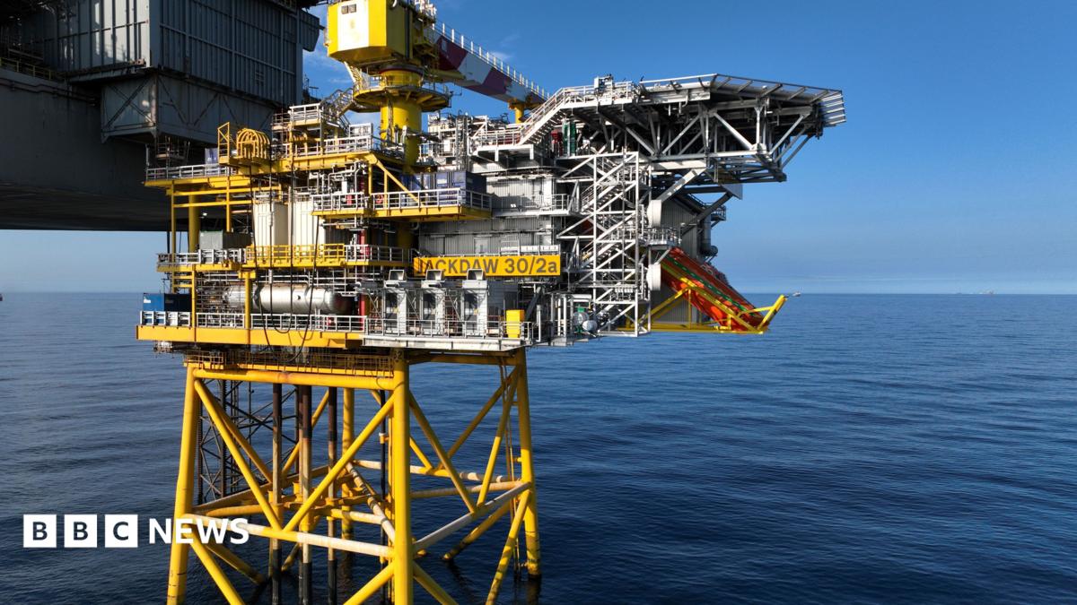 A mostly yellow-painted gas platform and allied grey rig stand in calm blue seas with a clear blue sky behind them. It is sunny.