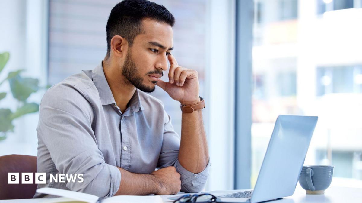 A man in a pale shirt sits at a laptop in an office