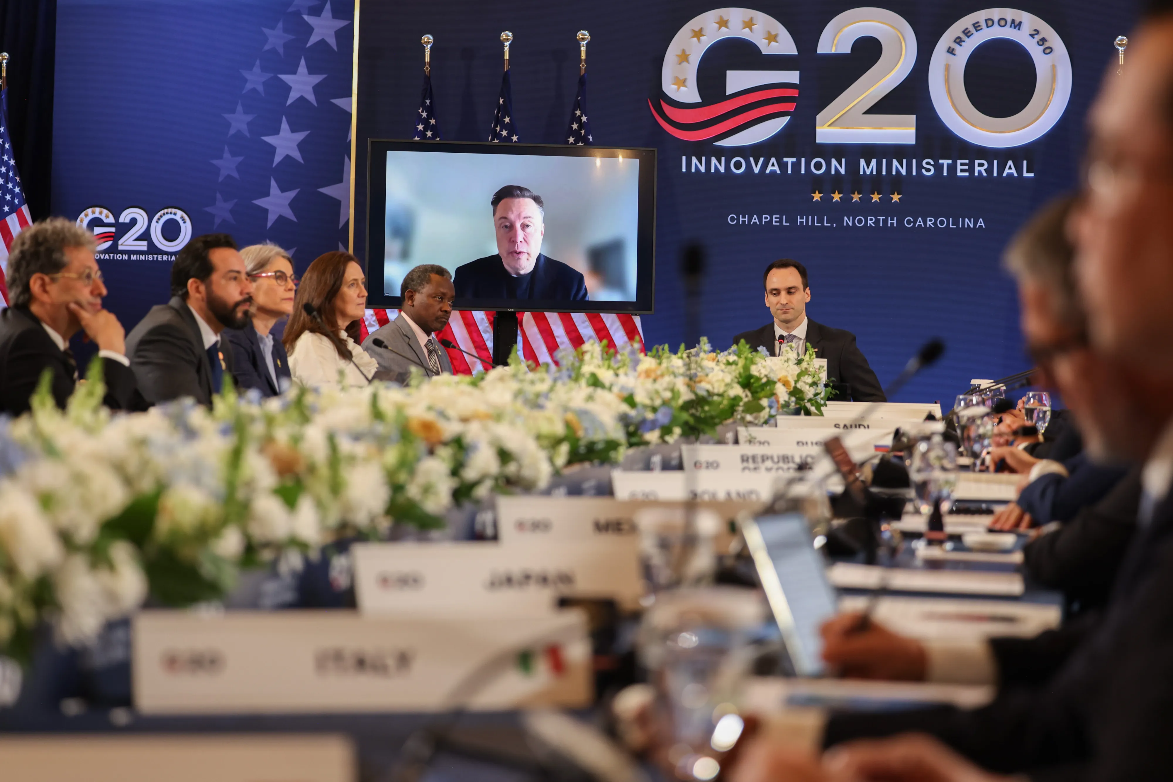A person on a livestream speaks to a long table in front of a G20 sign