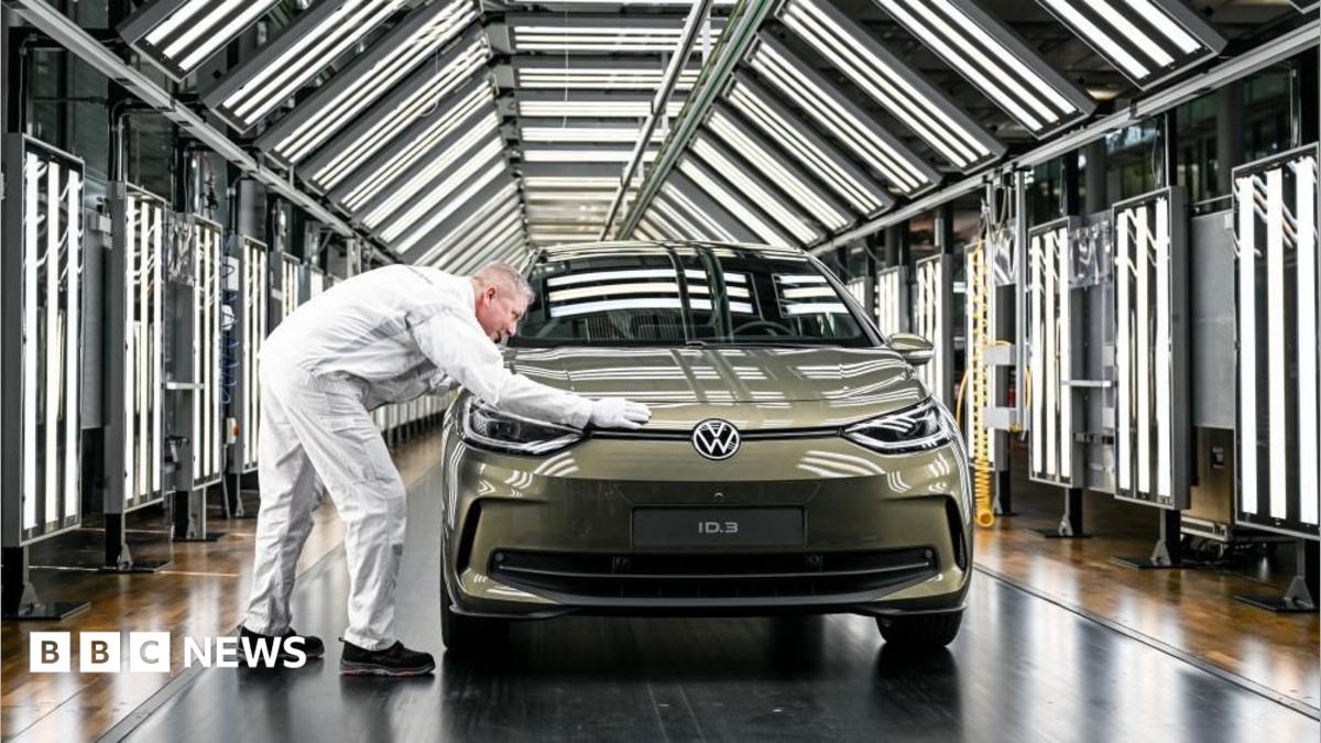 A Volkswagen employee presents a Volkswagen ID.3 car during a press presentation. He has his hand over a car, in a mossy green colour, parked in a tunnel full of white LED panels.